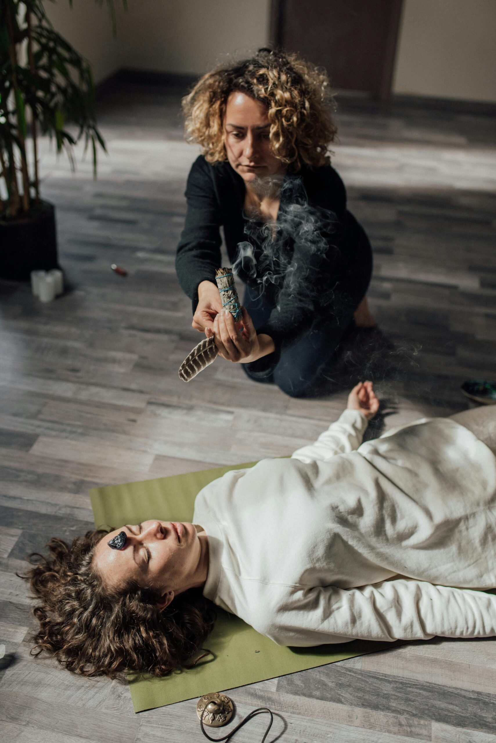 A Woman Holding a Feather and White Sage Beside a Woman Lying on Floor